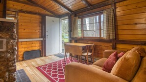 Interior acogedor de una cabaña con techo de madera, pared de piedra, zona de comedor con mesa y sillas, y un sofá con cojines.