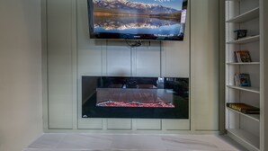 Un televisor de pantalla plana montado en la pared, con reflejo de montañas y lago, un televisor debajo, y una estantería con libros y un juego de mesa.
