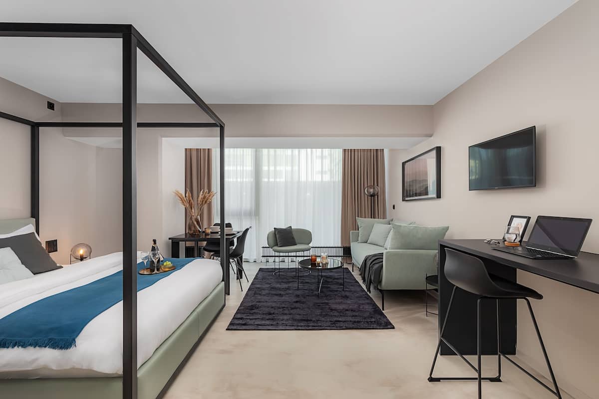Et moderne hotelværelse med himmelseng, skrivebord med en bærbar computer, en behagelig lænestol og et spiseområde med et bord og stole.