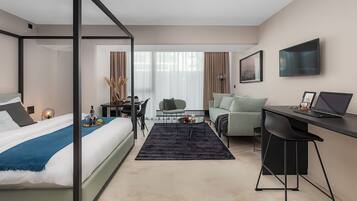 A modern hotel room with a four-poster bed, a desk with a laptop, a comfortable armchair, and a dining area with a table and chairs.
