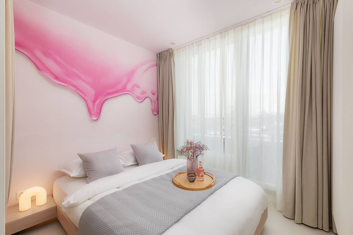 Et moderne hotelværelse med seng, et sengebord, gardiner ved et vindue og en væg med et abstrakt rosa design.