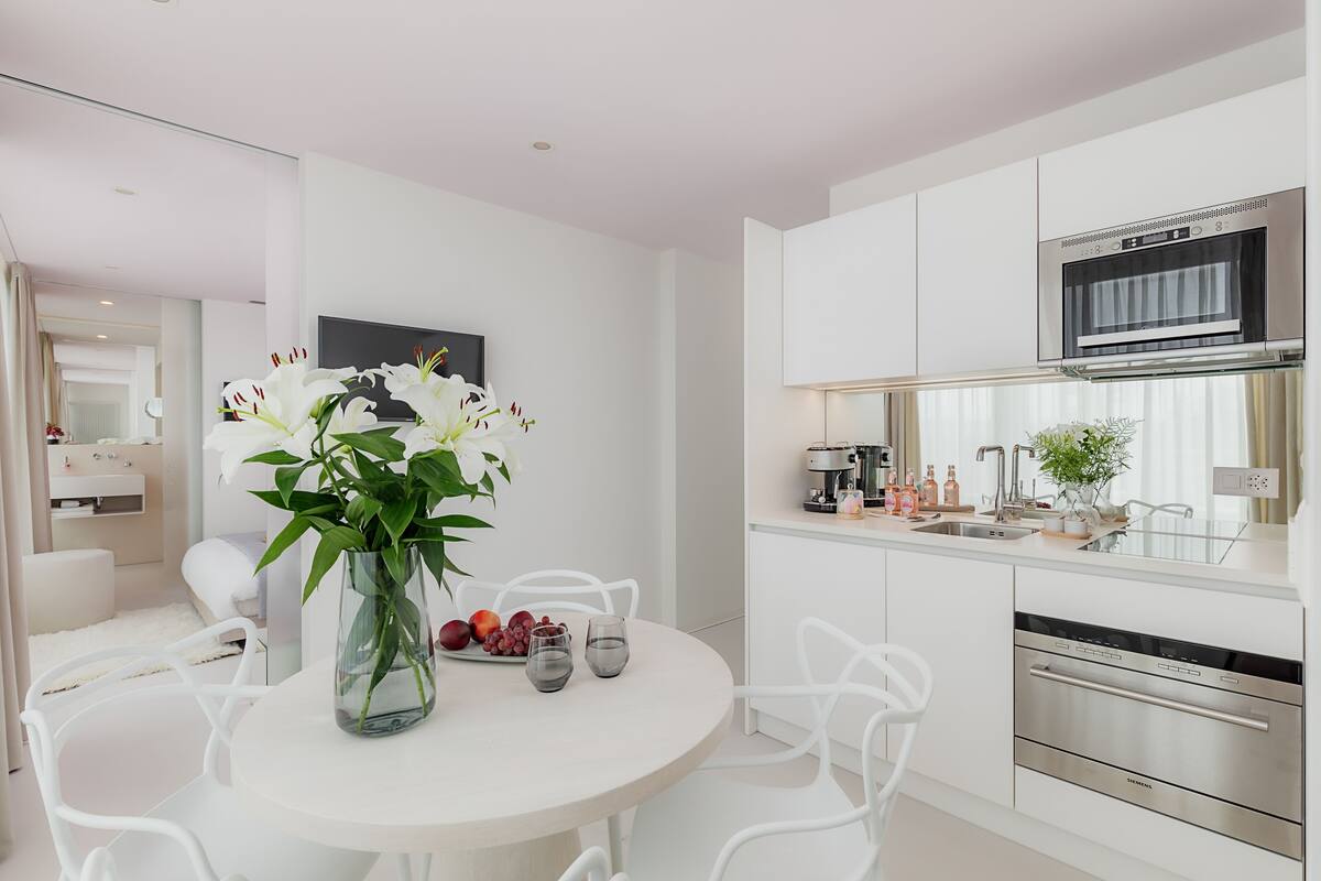Et moderne køkken med hvide skabe, indbygget ovn og mikrobølgeovn. Der er et spiseområde med et hvidt bord og stole, samt en vase med blomster på bordet.