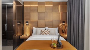 Una habitación de hotel moderna con cabecera de madera, cama con ropa blanca, un mesero con vasos y una botella, y un ventanal con cortinas.