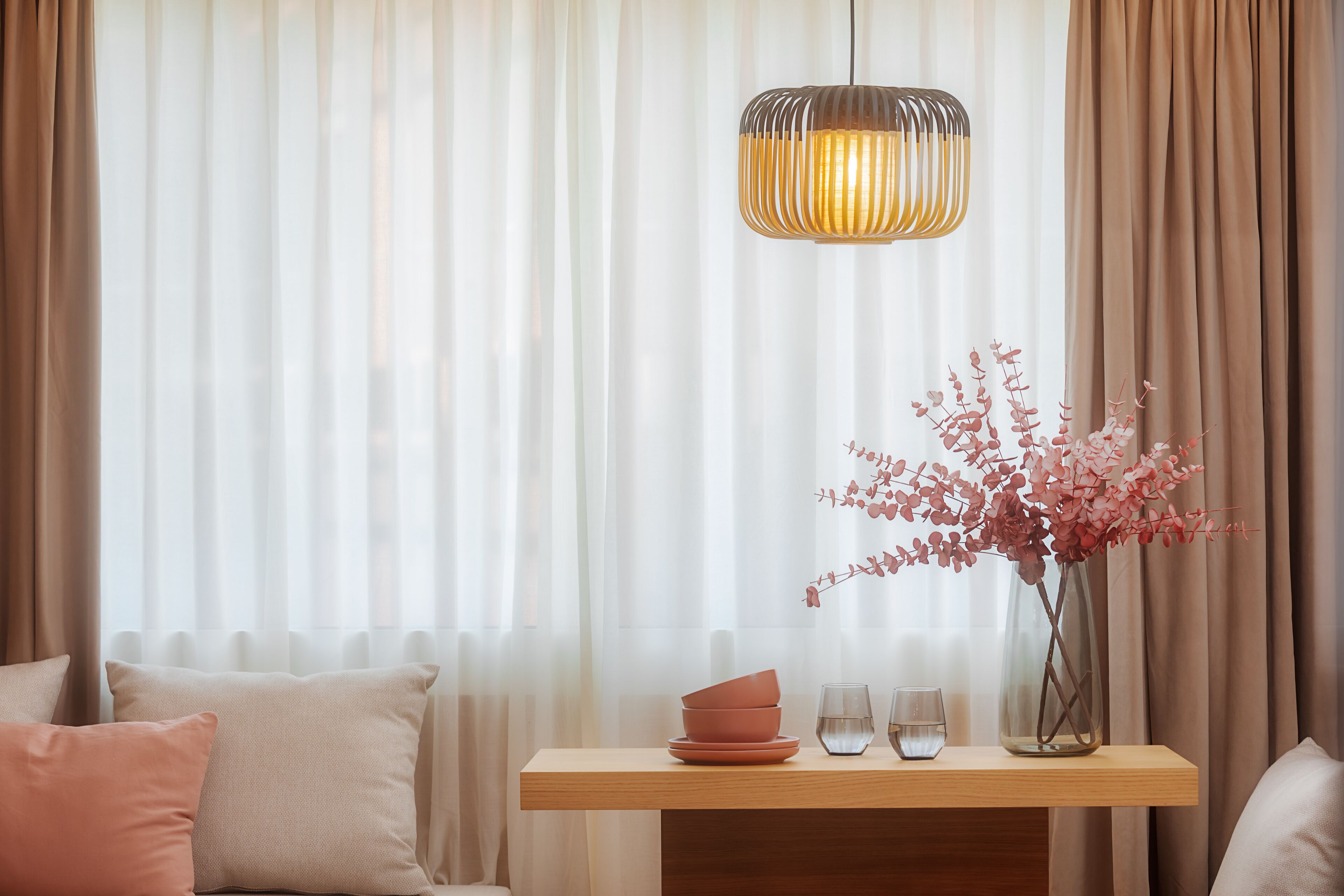 A room with a wooden table, a vase with pink flowers, and a hanging light fixture.