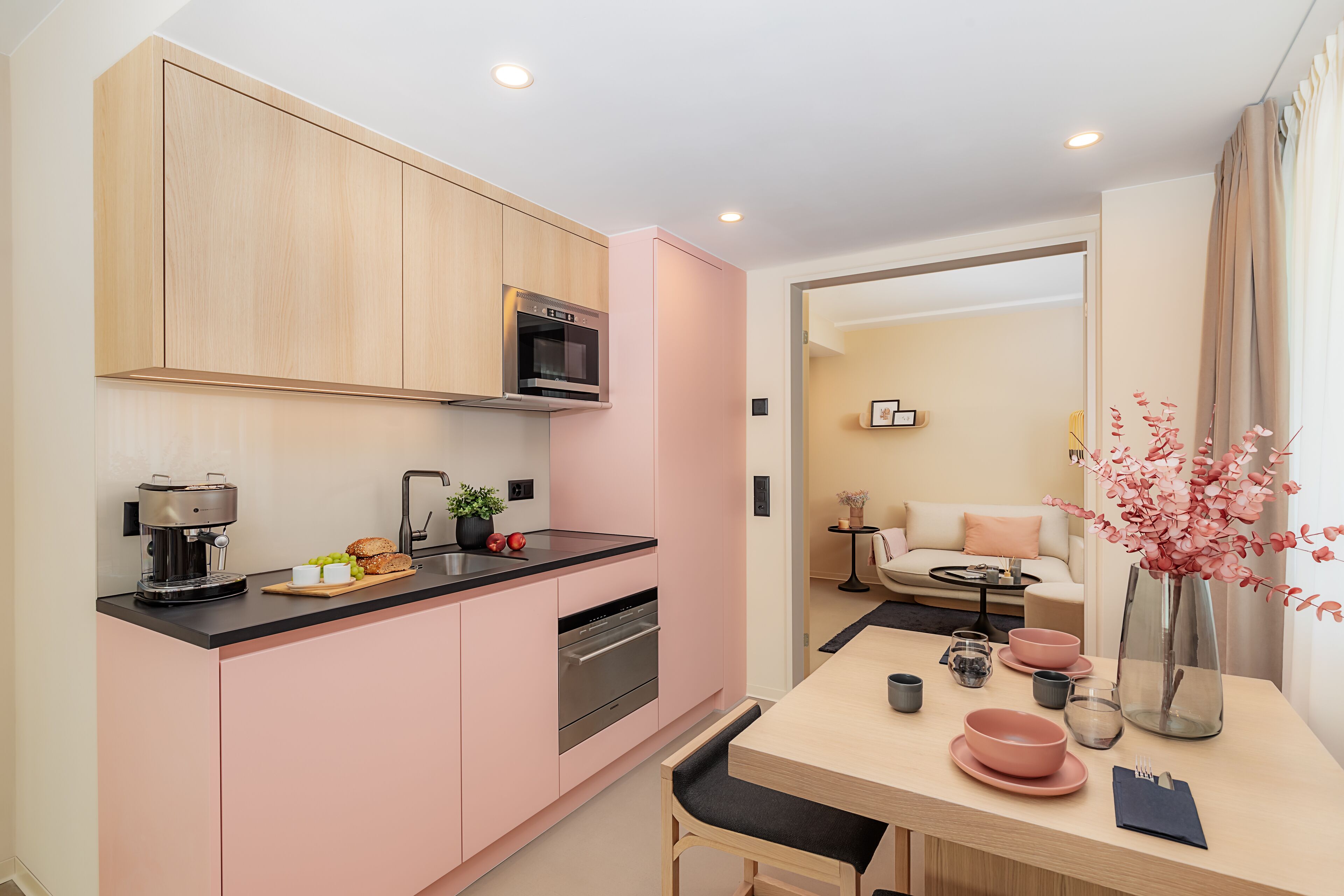 A modern kitchen with pink cabinets, a black countertop, and a dining area with pink bowls and a vase of flowers.