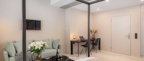 Un dormitorio moderno con una cama, un sofá, una mesa de centro con flores, un escritorio con una computadora portátil y un televisor en la pared.