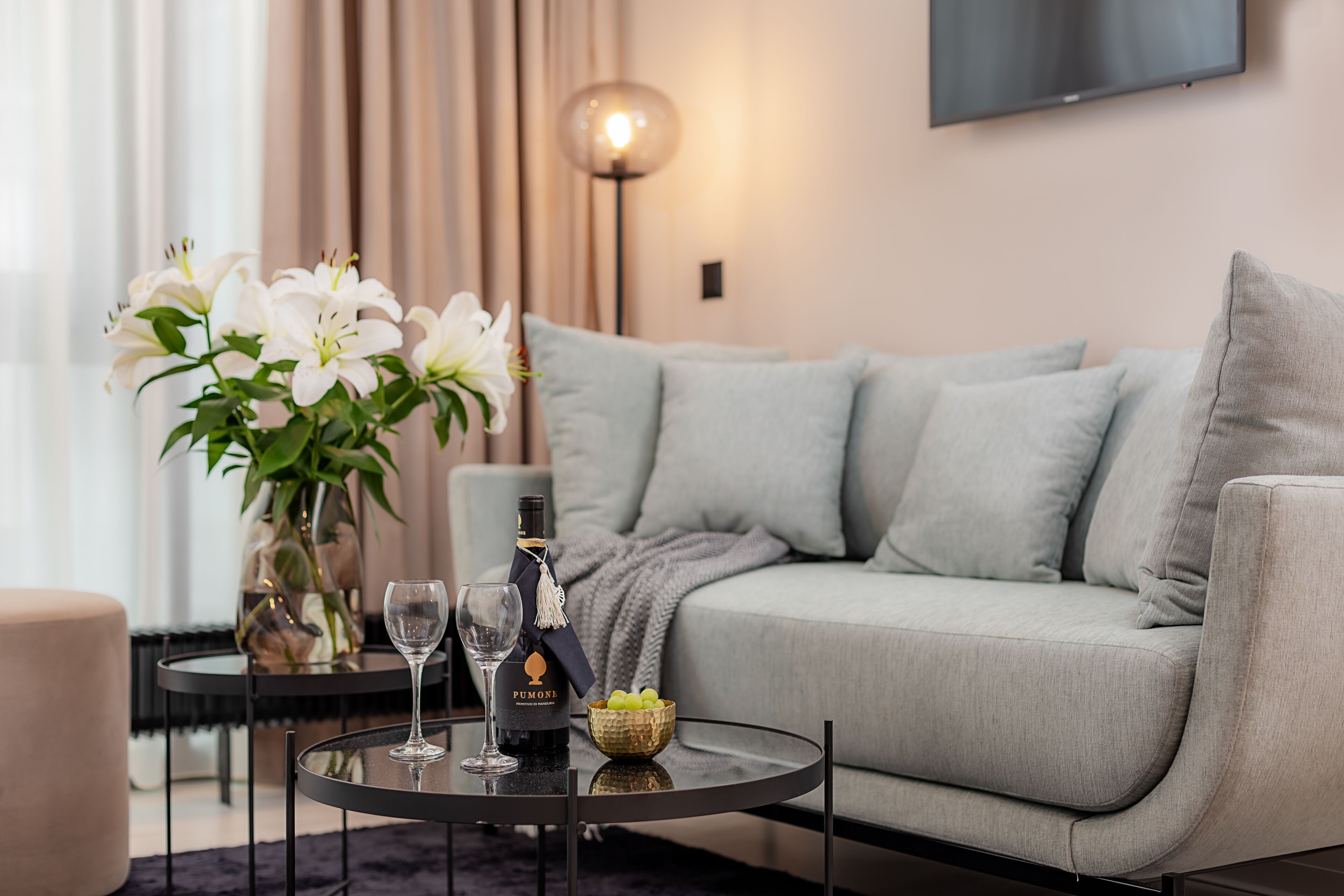 Un salon moderne avec un canapé gris, une table basse ronde en verre, un vase avec des fleurs blanches, une bouteille de vin et un saladier de fruits.
