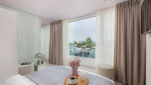 Una habitación de hotel moderna con una cama grande, una mesita de noche con una lámpara, una bandeja con una taza y un jarrón de flores, y vistas a un balcón con mobiliario exterior.