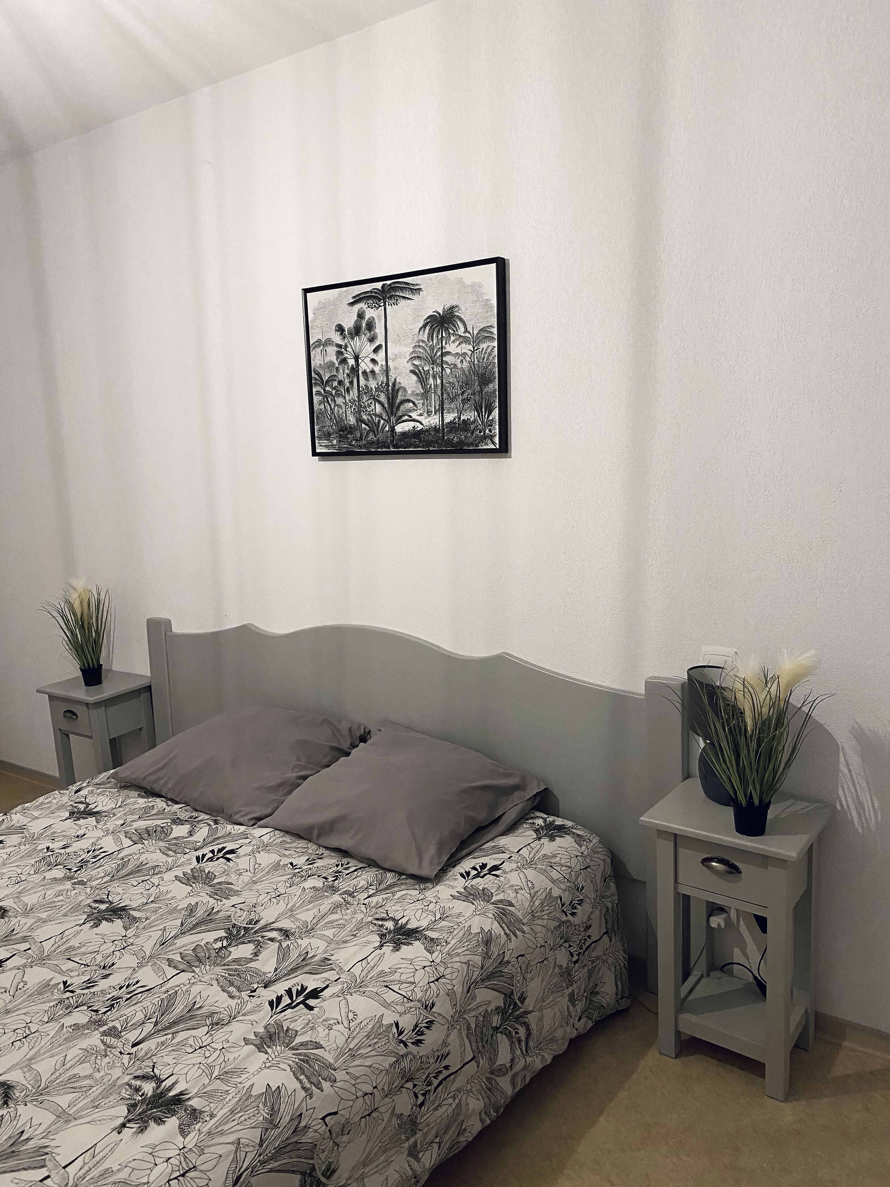 Une chambre à coucher avec un lit, une table de chevet, un tiroir, un tableau encadré et une plante en pot.