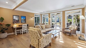 Ein geräumiges Wohnzimmer mit einer Couch, Sesseln, einem Couchtisch und einem Essbereich. Zwei große Fenster bieten Blick ins Freie.