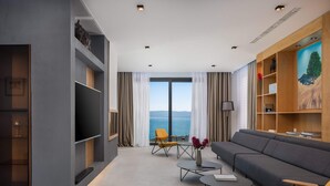 Una sala moderna con un televisor de pantalla plana grande, un sofá gris, sillas amarillas y vistas al mar a través de amplios ventanales.