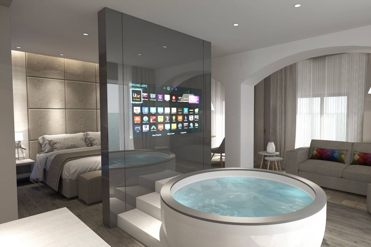 Una habitación de hotel moderna con un televisor grande montado en la pared que muestra diversas aplicaciones, una zona de estar acogedora con sofá y sillón, una mesita y una bañera de hidromasaje circular.