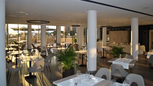 Un comedor amplio con varias mesas y sillas, ventanas grandes y plantas en macetas.