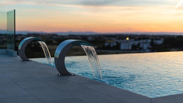 Piscina moderna al aire libre con dos fuentes y vistas a la ciudad al atardecer.