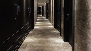 Un pasillo largo y estrecho con suelo de baldosas, bordeado por paredes y puertas oscuras, que conduce a una salida luminosa al final.