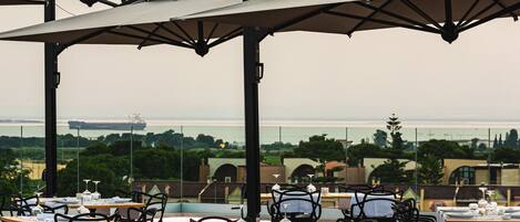 An outdoor dining area with tables and chairs, a view of the sea, and a ship in the distance.