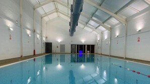 En indendørs swimmingpool med klart, blåt vand, fliset gulv og et hvælvet loft med synlige bjælker.