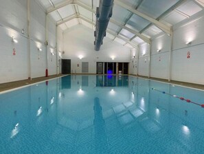 An indoor swimming pool with clear blue water, a tiled floor, and a high ceiling with visible structural beams.