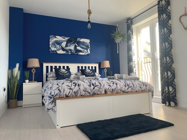 Une chambre à coucher comprenant un lit, des tables de chevet, un luminaire suspendu, une fenêtre avec des rideaux et un tableau mural.