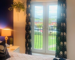 A bedroom with a bed, a bedside lamp, a potted plant, and a view of a green lawn and houses through a sliding door. - Sandy Feet 1st floor apartment with open plan living on The Bay, Filey (Primrose Valley)
