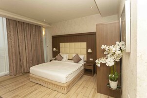 Una habitación de hotel con una cama grande, cabecera de madera, mesitas de noche con lámparas, una orquídea en maceta y un amplio ventanal con cortinas.