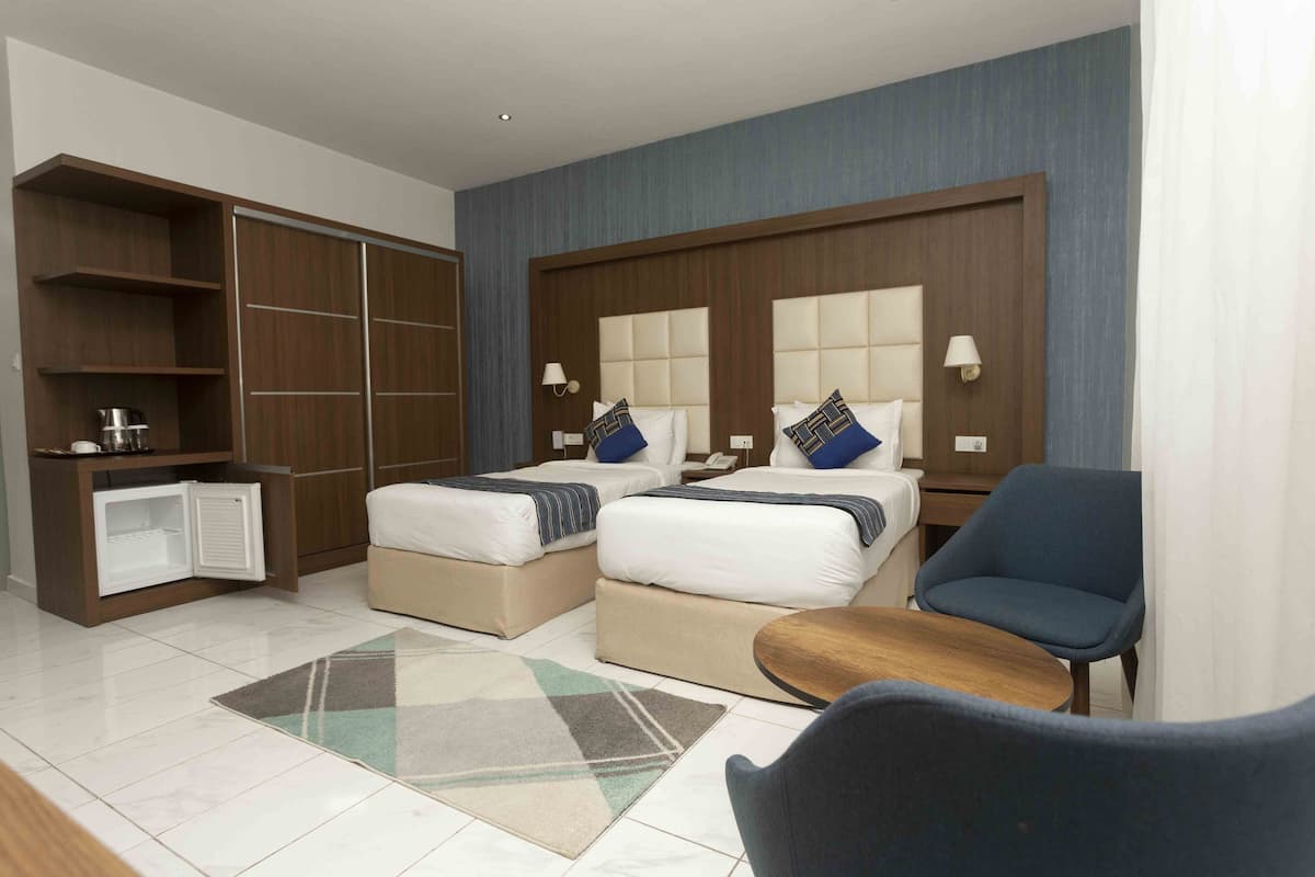 A hotel room with two beds, a small seating area, a wardrobe, and a mini-fridge.