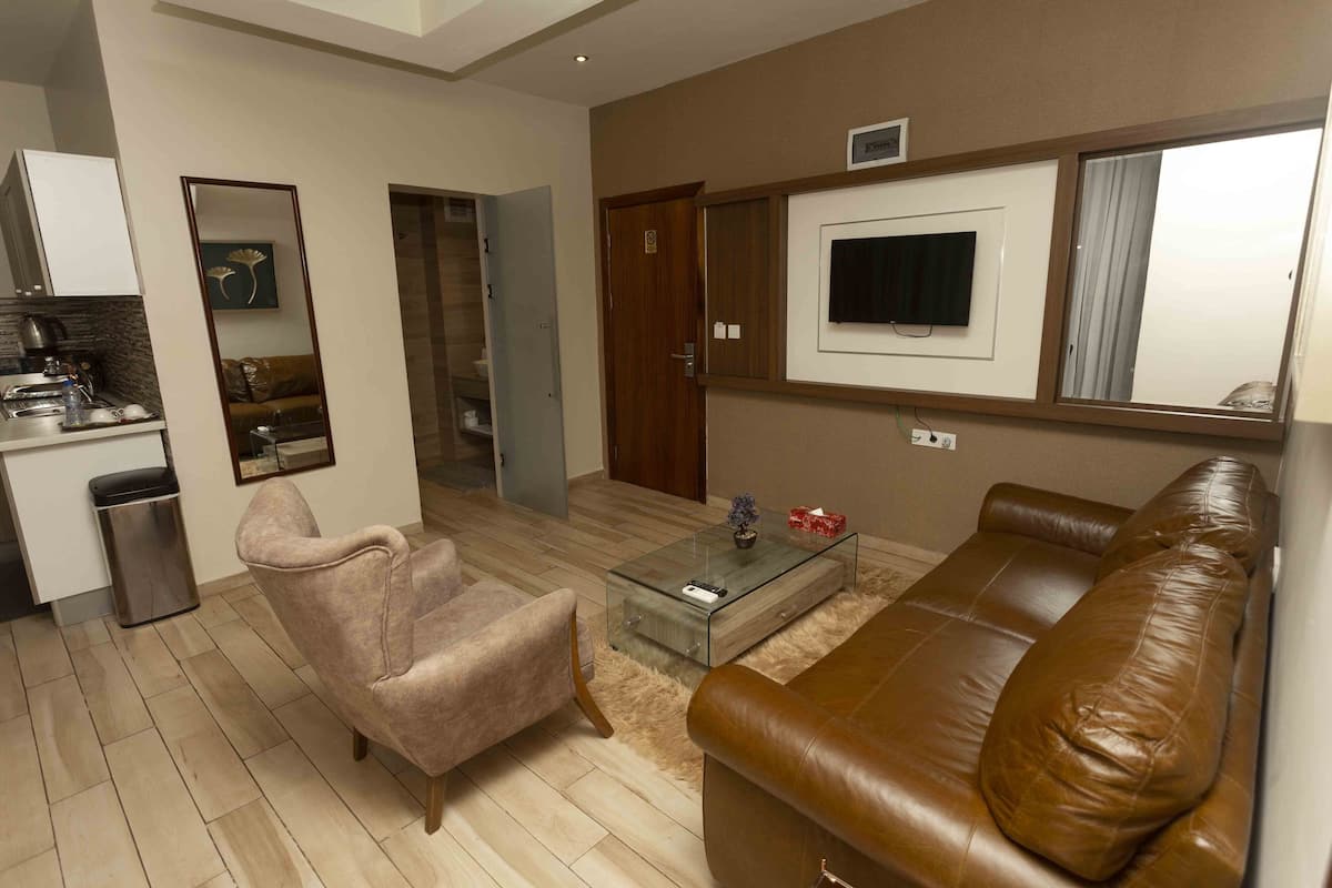 A modern living room with a brown leather sofa, a glass coffee table, a beige armchair, a kitchen area with a sink and cabinets, and a TV mounted on the wall.