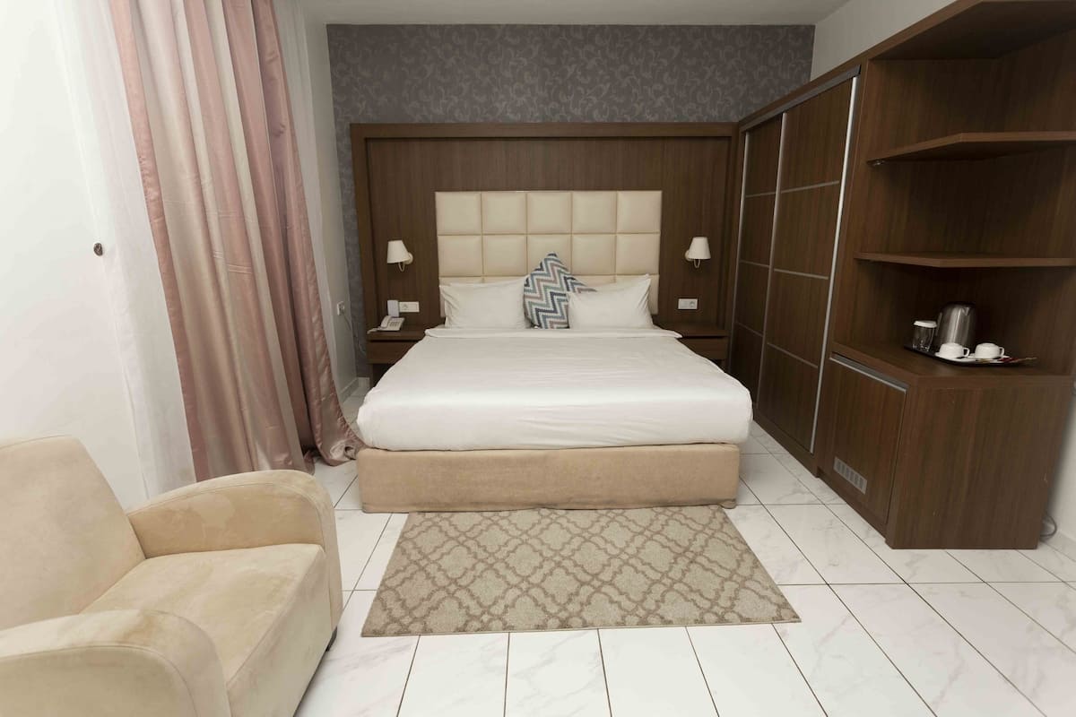 A hotel room with a large bed, bedside lamps, a wooden wardrobe, a beige sofa, and a patterned rug.