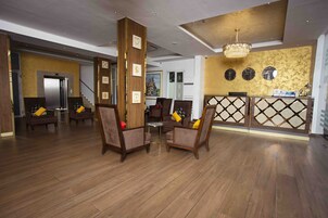 Un hall con suelo de madera, dos sillones con cojines amarillos, un mostrador de recepción y una araña.
