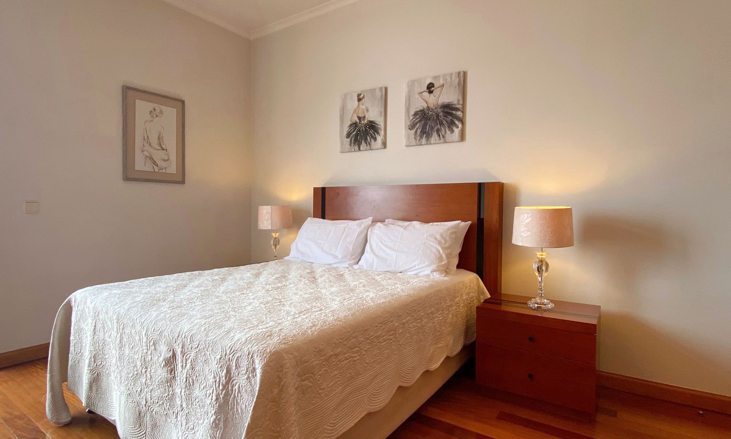Un lit bien fait, recouvert d’une couvre-lit blanche, avec deux lampes de chevet et une table de chevet en bois dotée d’un tiroir.