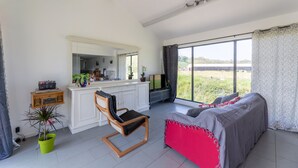 A spacious room with a large window, a bed, a chair, a kitchen area, and a potted plant.