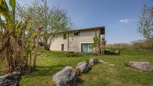 A stone house with a tiled roof, a small patio, and a grassy yard with large rocks and banana trees.