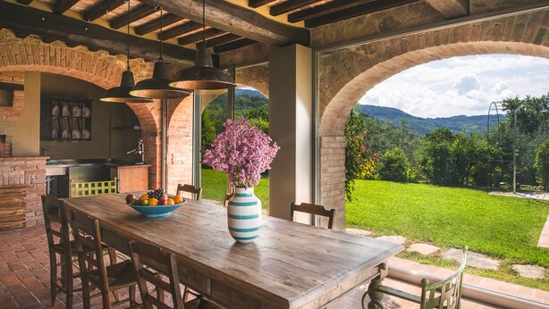 An outdoor dining area with a rustic brick wall, a wooden table, and a bowl of fruit. - Private villa with pool with panoramic view inside organic wine resort (Arezzo)