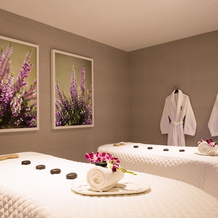 A spa room with two beds, each with a rolled towel and a flower arrangement, and two robes hanging on the wall.