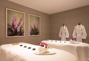 A spa room with two beds, each with a rolled towel and a flower arrangement, and two robes hanging on the wall. - The Equestrian Hotel (Ocala)