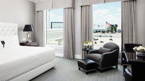 A hotel room with a large bed, a chair, a small table with flowers, and a view of a bridge and flags. - The Equestrian Hotel (Ocala)