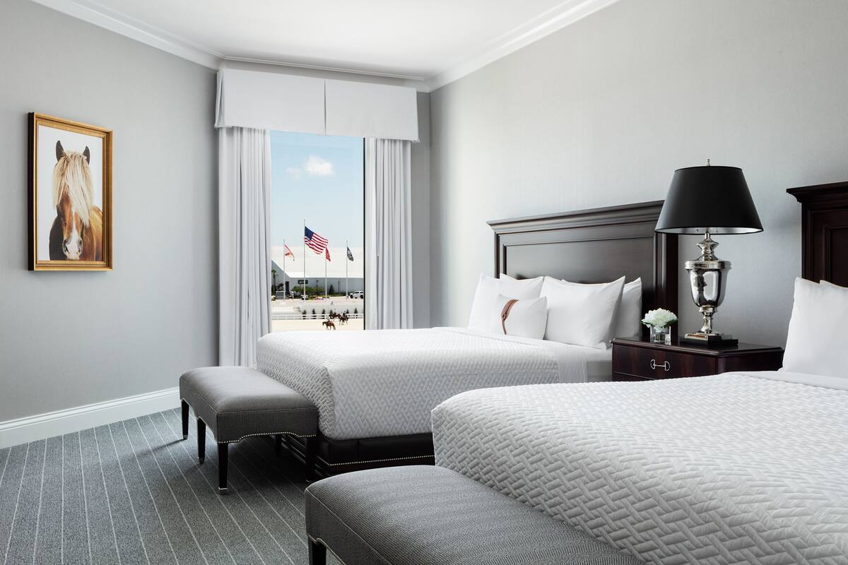 A hotel room with two beds, a nightstand, a lamp, a framed picture, and a view of a street with flags.