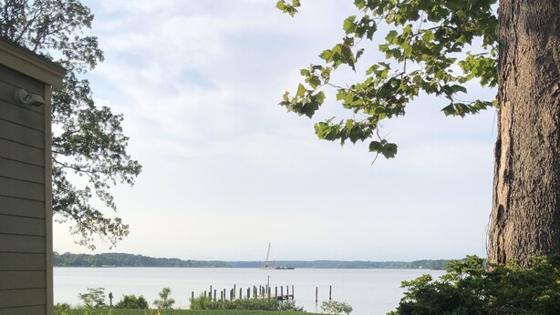 A grassy yard with a wooden deck, a large tree, and a view of a calm body of water with a distant shoreline. - Salty Air, Sandy Toes - Quiet Escape w/ Kayaks & Sandy Beach - 2BD waterfront (Heathsville)