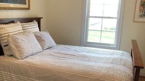 A bedroom with a large bed, a window with blinds, a colorful rug, and a painting on the wall. - Salty Air, Sandy Toes - Quiet Escape w/ Kayaks & Sandy Beach - 2BD waterfront (Heathsville)