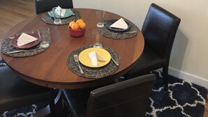 A dining area with a round table set for four, surrounded by black leather chairs. A framed seascape painting hangs on the wall above the table. - Salty Air, Sandy Toes - Quiet Escape w/ Kayaks & Sandy Beach - 2BD waterfront (Heathsville)