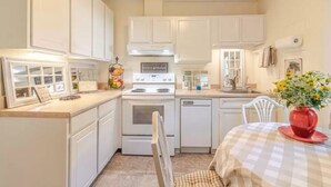 A compact kitchen with white cabinets, a stove, and a dishwasher. There is a dining table with a checkered tablecloth and a red vase with flowers. - Salty Air, Sandy Toes - Quiet Escape w/ Kayaks & Sandy Beach - 2BD waterfront (Heathsville)