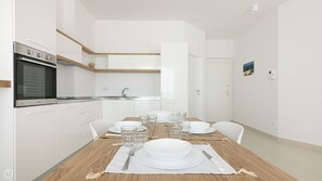 A modern kitchen with white cabinets, a built-in oven, and a wooden dining table set for four with plates, glasses, and cutlery.