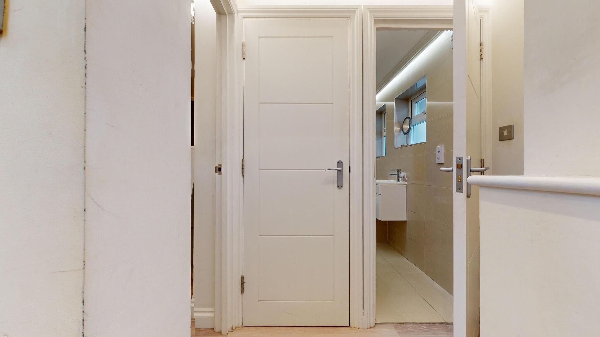 A hallway with a white door, a bathroom with a sink and mirror, and a light switch on the wall.