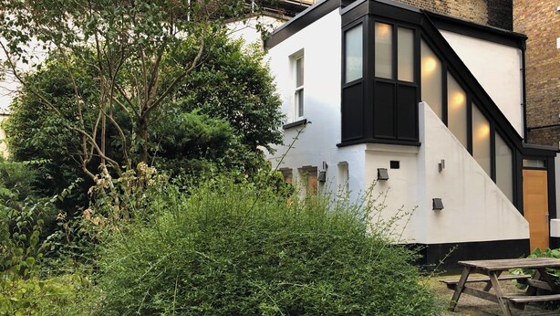 Een modern huis met twee verdiepingen, een witte gevel en zwarte accenten, omgeven door weelderige begroeiing.