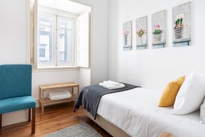 A bedroom with a bed, a blue chair, a wooden bench, and a window with white shutters.