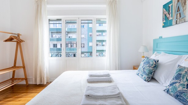 A neatly made bed with white linens and blue patterned pillows, a wooden shelf, and a window with a view of a building. - Casa da Avenida III (Lisbon)