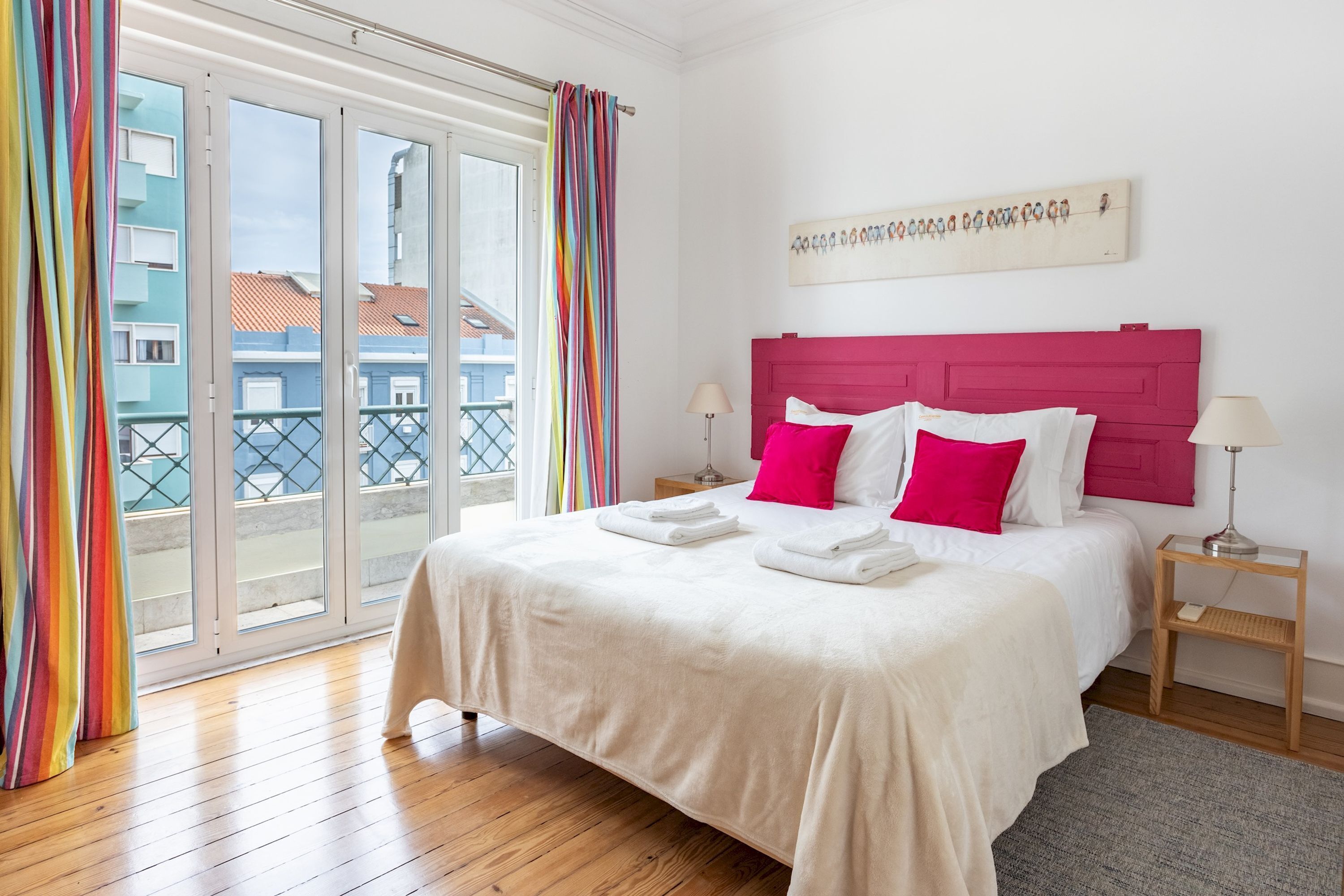 A hotel room with a large bed, a balcony with colorful curtains, a wooden floor, and a view of buildings outside.