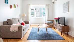 Una sala de estar con sofá, una mesa de centro, televisor y ventana con cortinas.