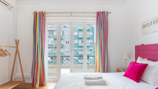 Habitación de hotel con cama, perchero de madera, ventana con cortinas y vista a un edificio.