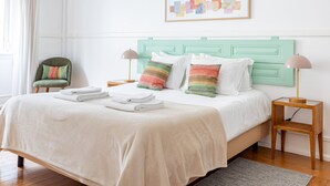Una cama bien hecha con edredón blanco, dos almohadas y toallas dobladas. Hay una silla verde y una mesita de noche con una lámpara.